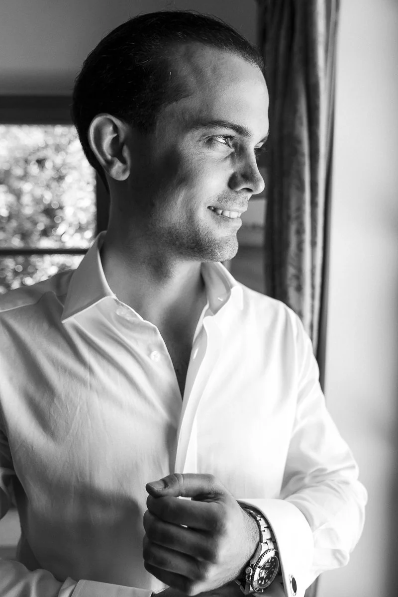 A black and white photo of a man with slicked-back hair wearing a white dress shirt and wristwatch, looking out a window with a slight smile.