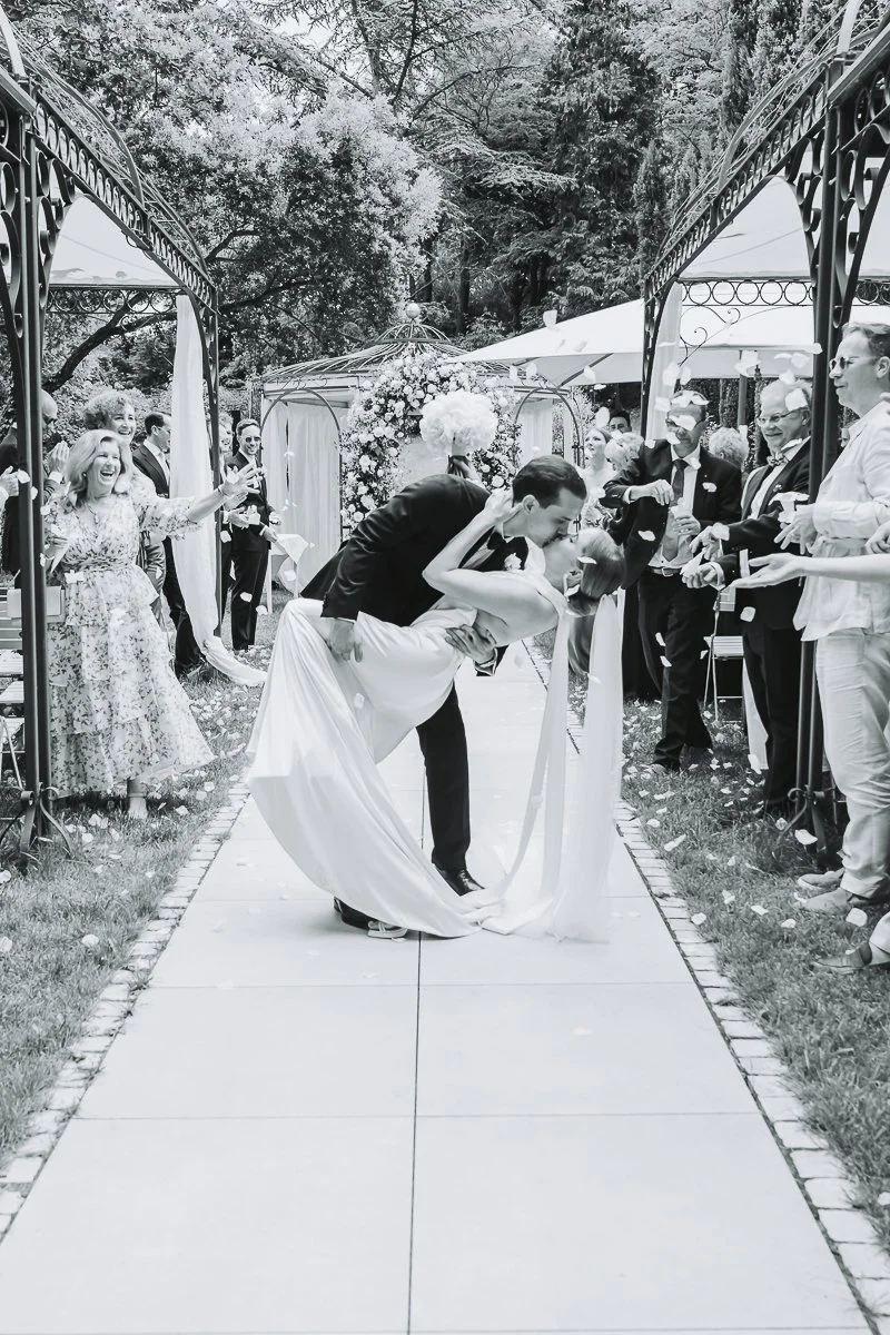 A couple kissing during their wedding ceremony outdoors, surrounded by friends and family, with a decorative floral arch in the background.