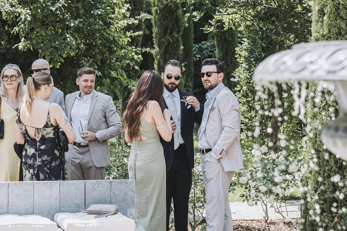 A group of people dressed in formal attire gathering outdoors in a garden with green trees and bushes.