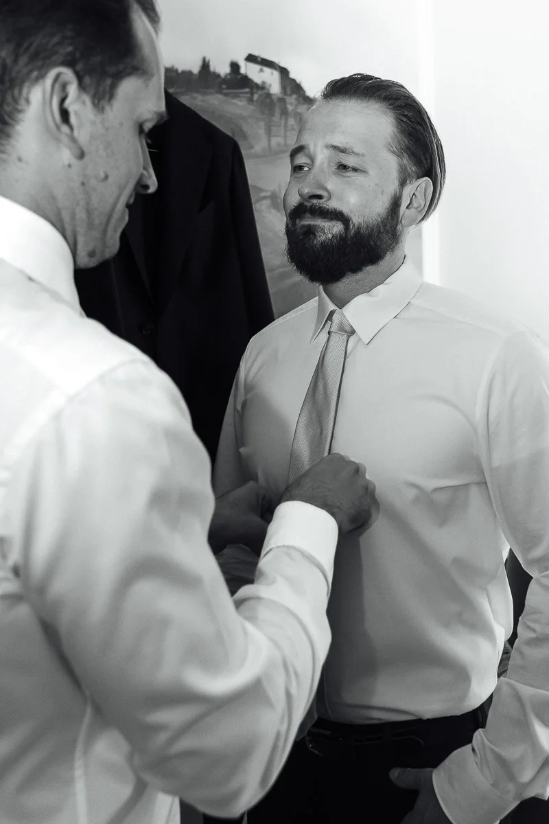 Two men in white shirts and ties are having a moment, with one pinning a boutonniere on the other.