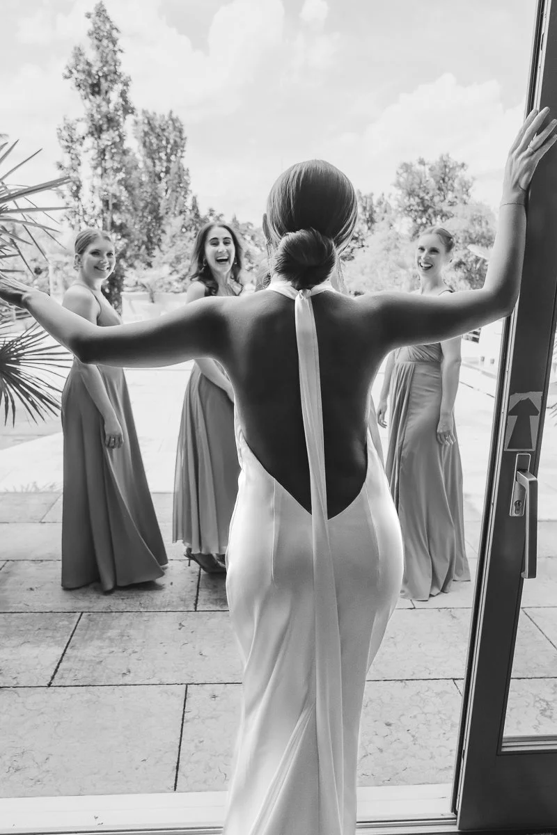 A bride in a backless wedding dress with a halter neck, facing her bridesmaids outside a building, all smiling.