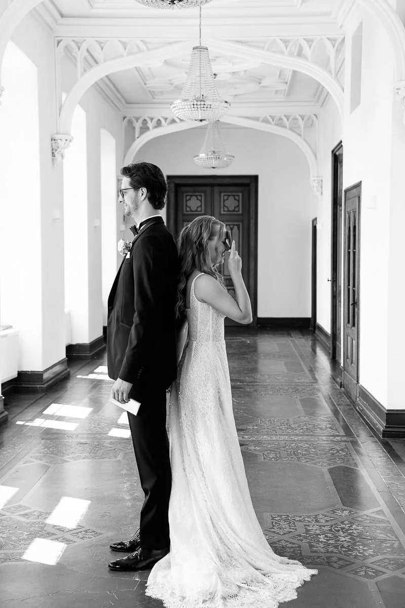 First Look: Braut tritt hinter den Bräutigam im Schlossflur, schwarz-weiß