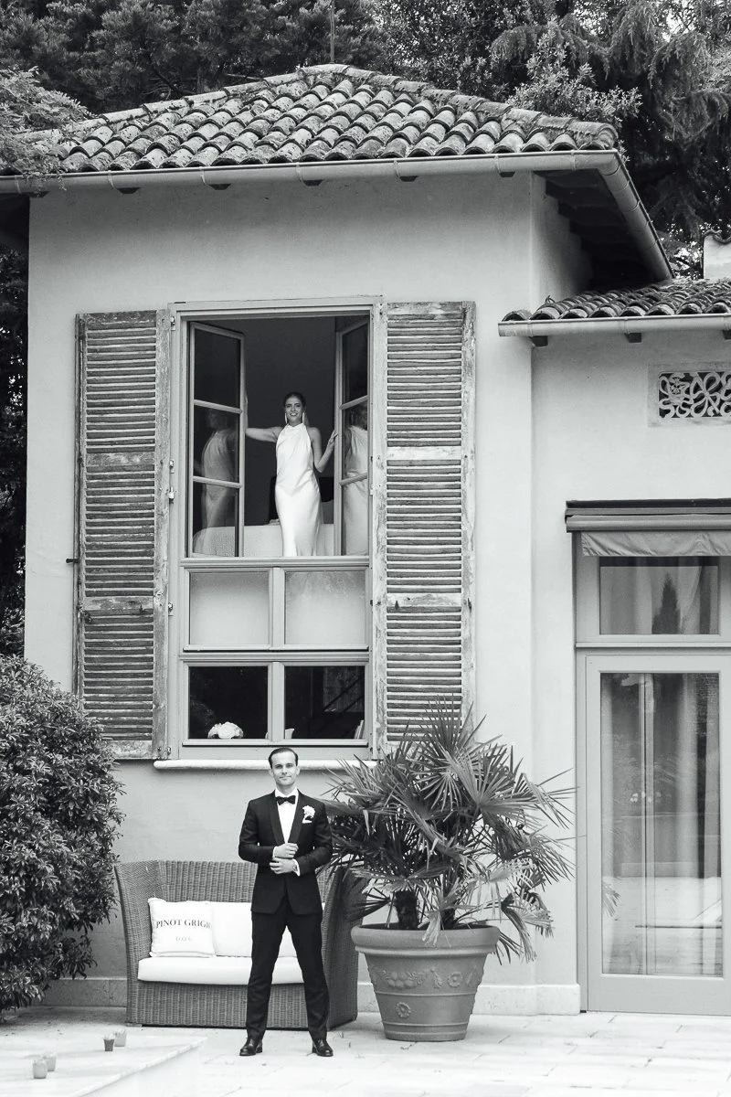A man in a tuxedo standing outside a house near a large potted plant with a couch and pillow nearby. A woman in a white dress is looking out from an open window above.