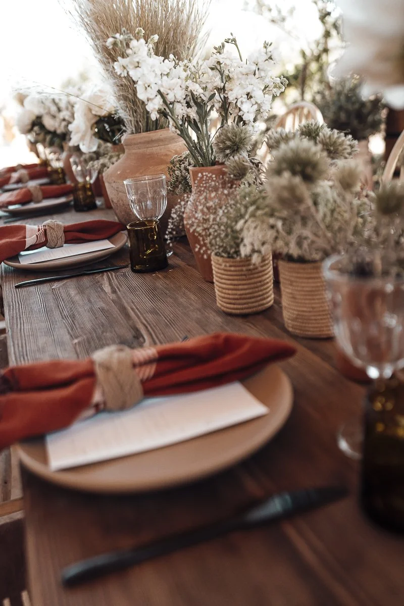 A rustic dining table decorated with numerous white and beige flowers in vases, set for a meal with plates, silverware, and glasses, creating a warm and inviting atmosphere.