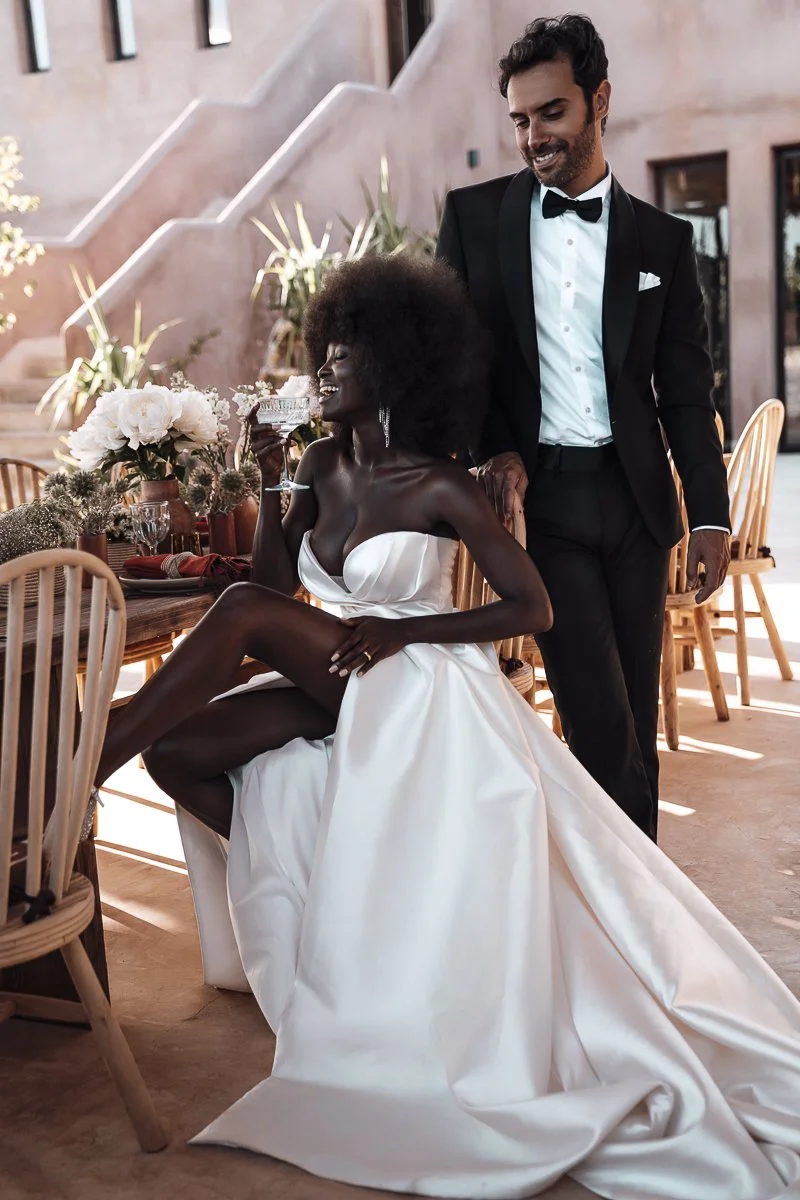 A bride in a white wedding dress sitting on a chair, holding a glass, and smiling, with a groom in a tuxedo standing beside her at a wedding reception outdoors.