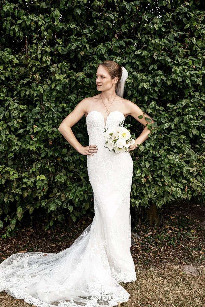 Braut im trägerlosen Brautkleid mit Brautstrauß vor grüner Hecke