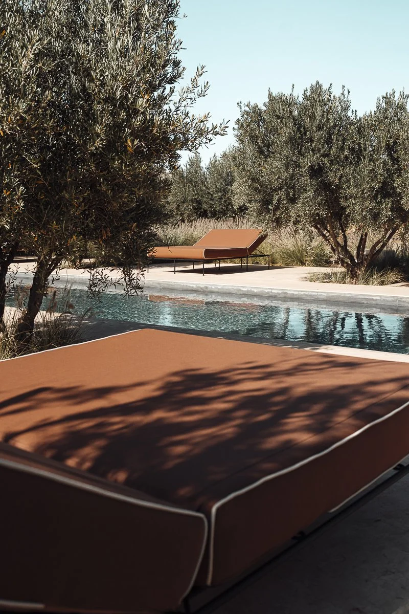 Outdoor poolside area with brown lounge chairs and green trees under a clear blue sky.