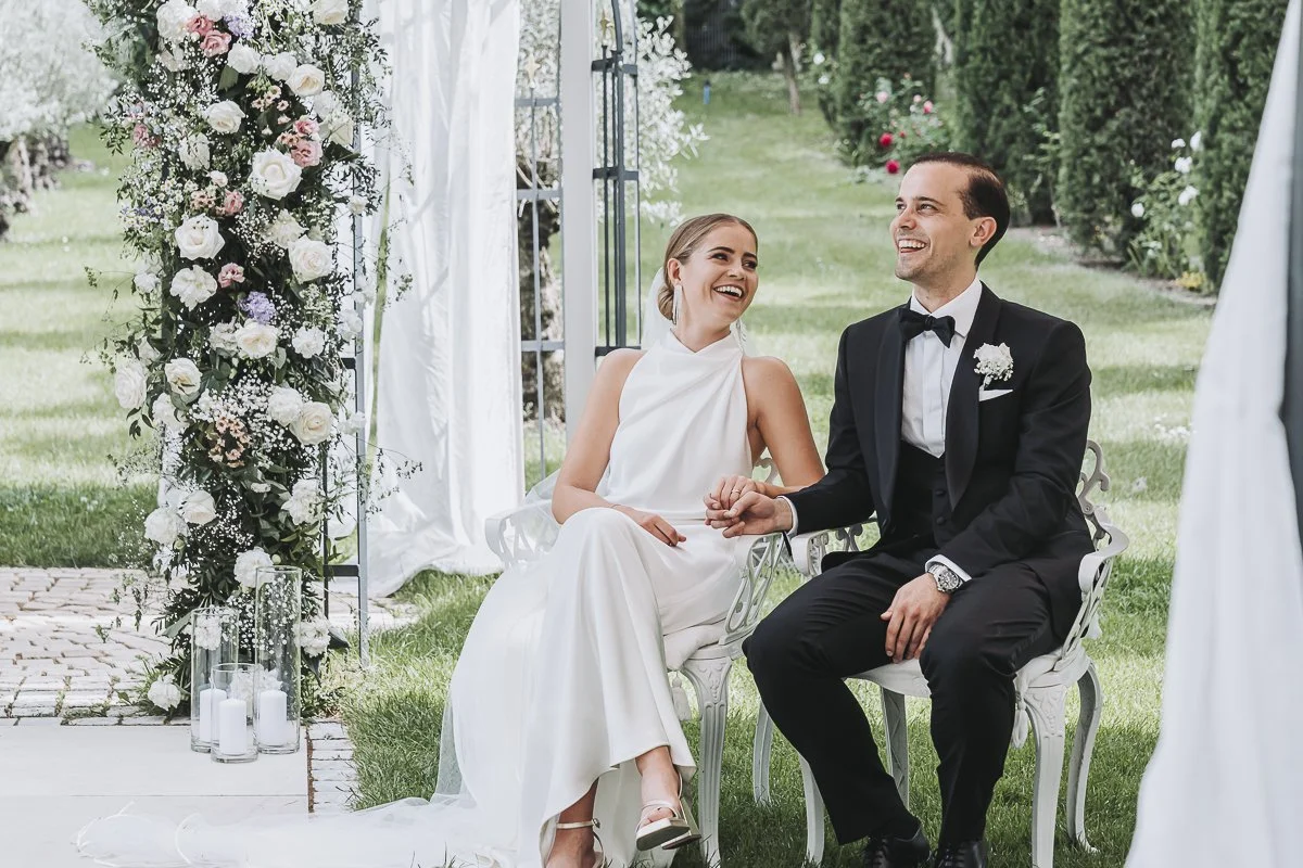 A bride and groom sitting on a white bench outdoors at their wedding, holding hands and smiling at each other. The bride is wearing a white sleeveless wedding dress, and the groom is in a black tuxedo with a bow tie. Behind them is a floral arch deco