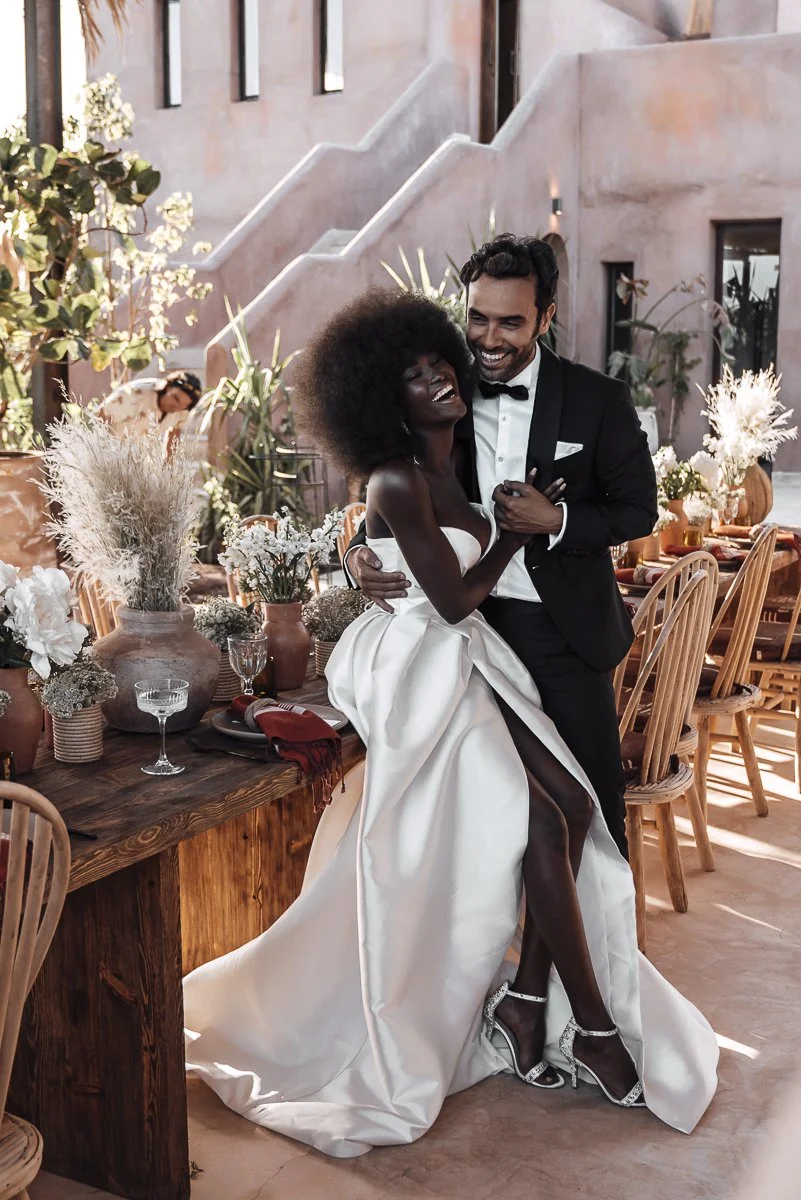 A joyful couple in wedding attire dancing at their wedding reception, with guests and decorated tables in the background.