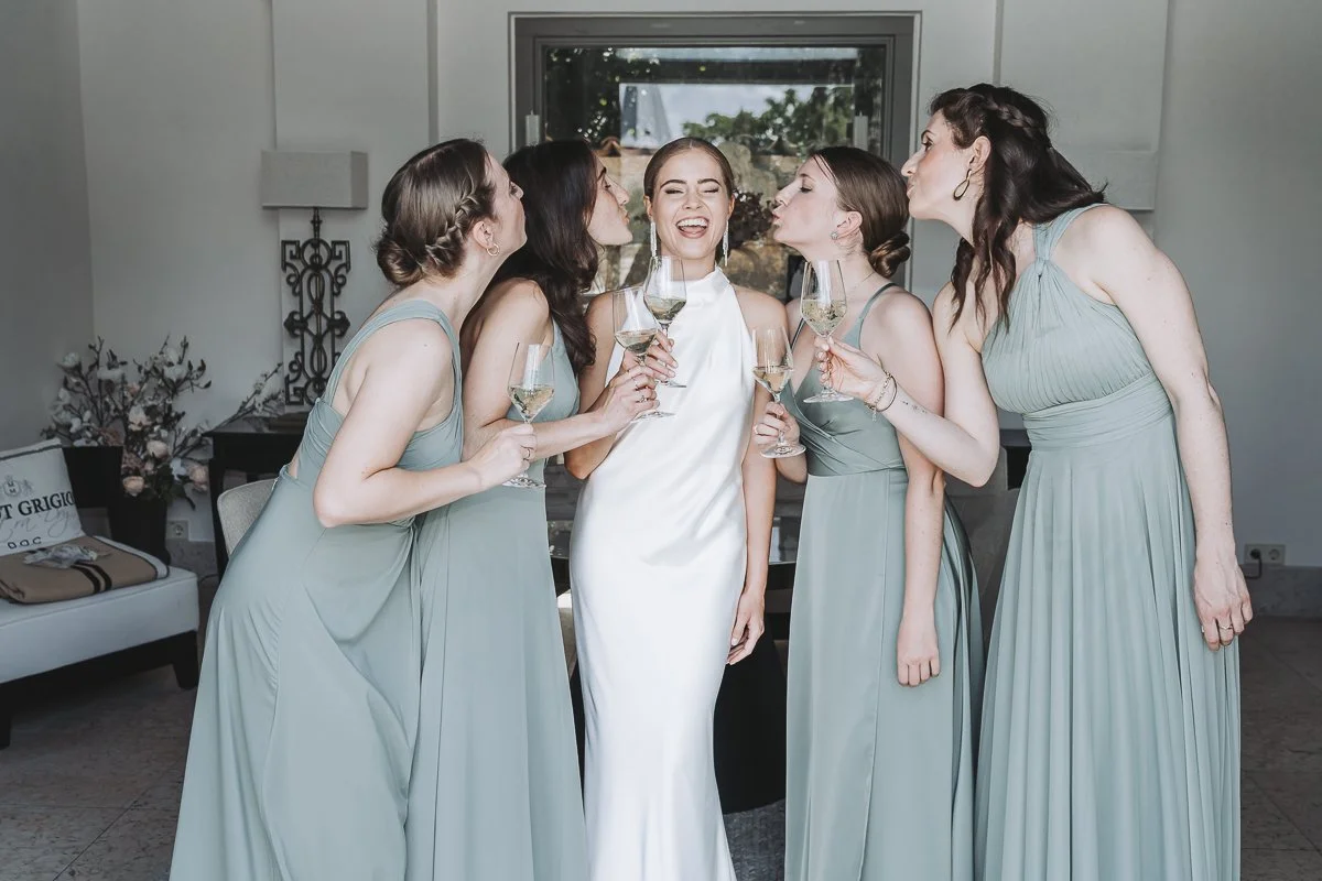 A bride in a white dress surrounded by five bridesmaids in matching light blue dresses, all holding glasses of white wine and appearing to kiss the bride on the cheeks.