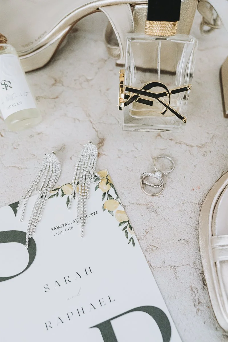 Jewelry, a perfume bottle, and a wedding invitation placed on a light-colored marble surface.
