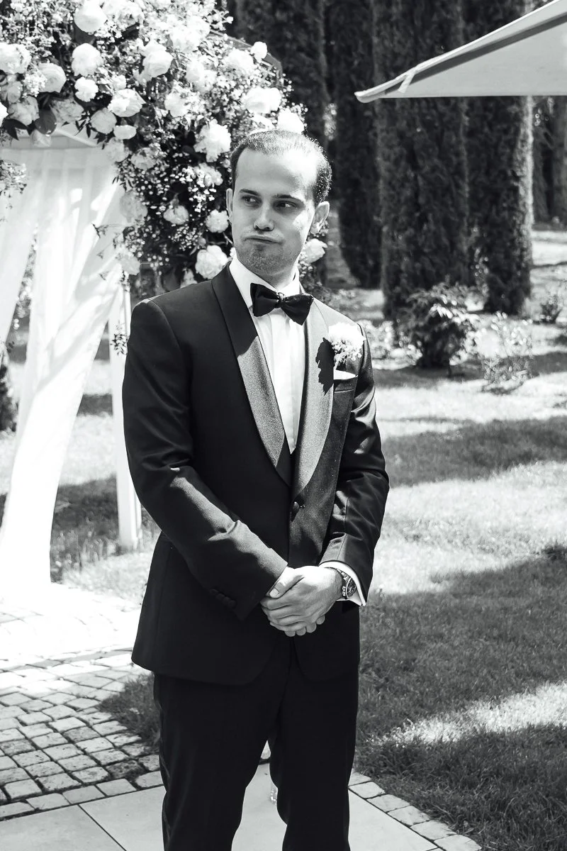 A man in a tuxedo with a bow tie, standing outdoors in front of a floral arch, during a formal event or wedding.
