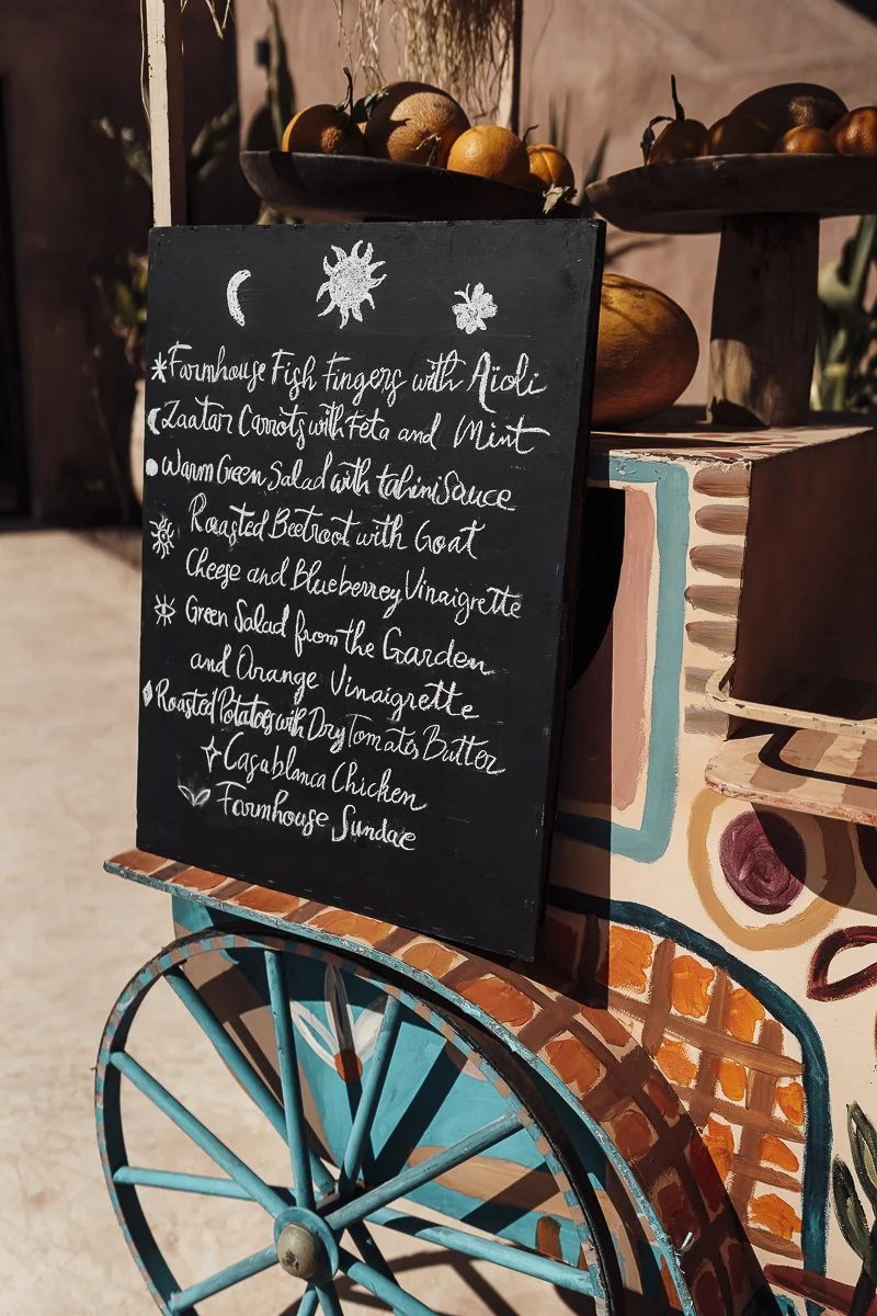 A black chalkboard sign with a menu written in white cursive, placed next to a colorful painted cart with a large blue wheel. The menu includes fish fingers with aioli, a salad, roast beetroot, a green garden salad, roasted potatoes, chicken, and a s