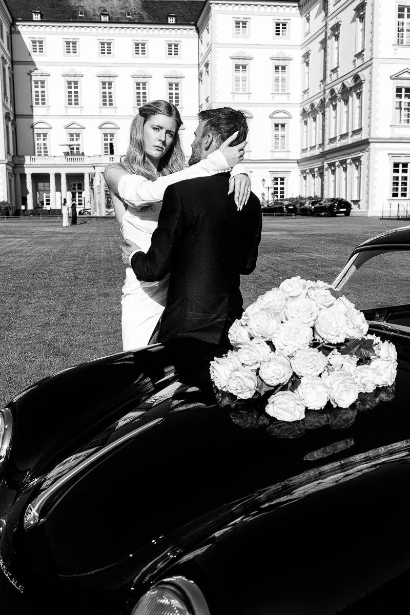 Brautpaar am schwarzen Porsche mit weißem Rosenschmuck, schwarz-weiß
