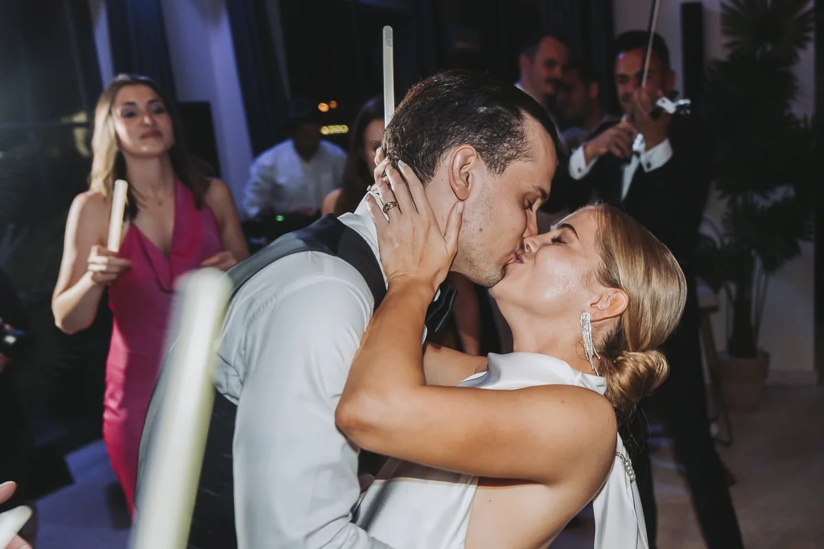A couple kissing at a celebration with guests in the background, some holding candles and playing violin.
