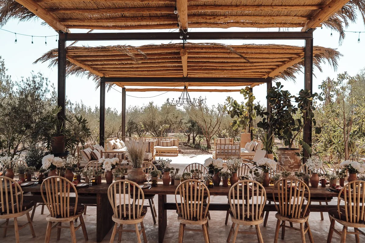 Outdoor party setup with a long wooden table decorated with flowers in vases, surrounded by wooden chairs, in a rustic patio area with a thatched roof and a view of trees in the background.