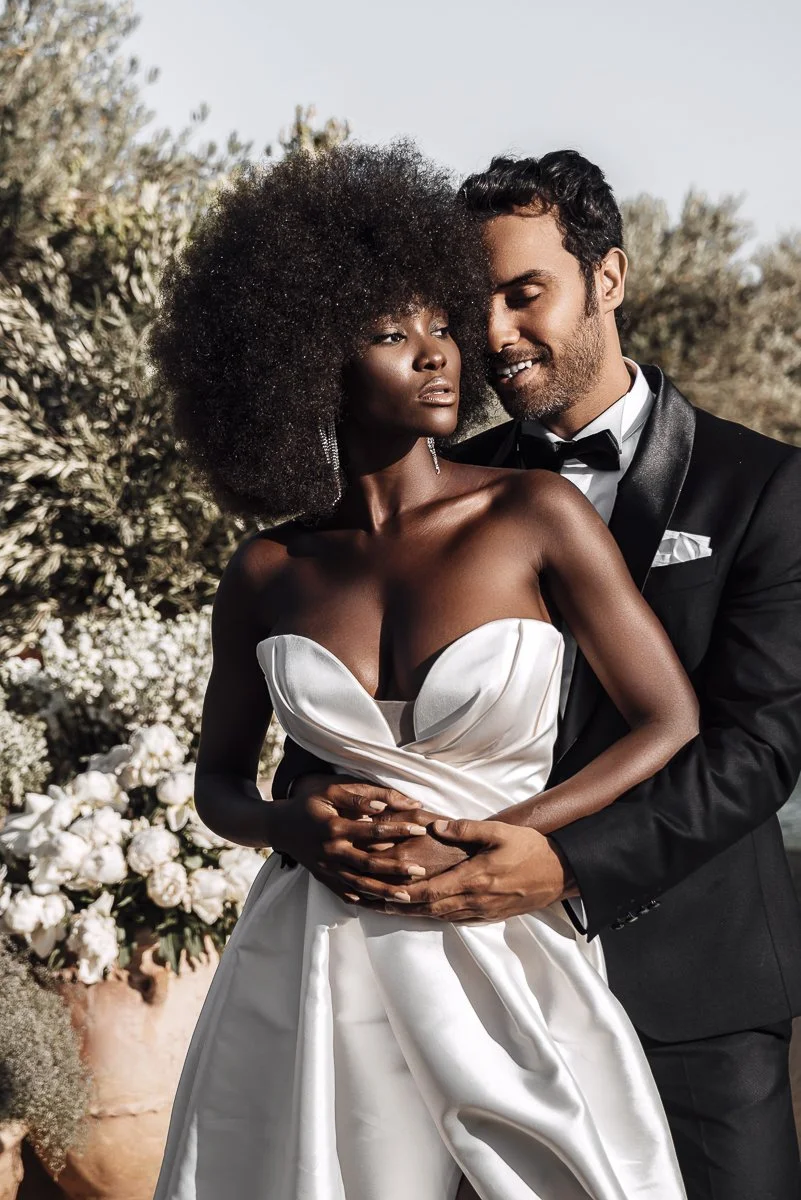 A couple dressed in wedding attire, with the woman wearing a strapless white gown and the man in a black tuxedo, holding each other outdoors near flowers.