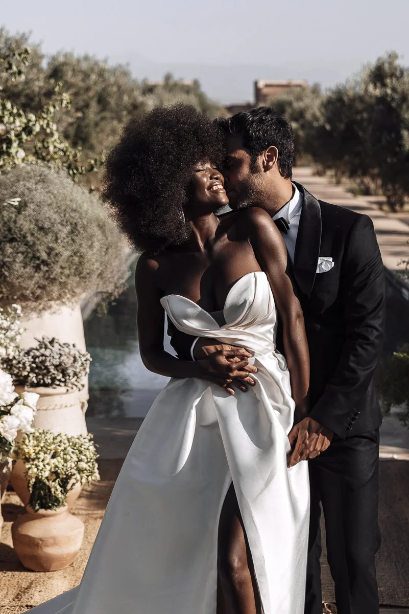A newlywed couple sharing a joyful moment outdoors. The woman is wearing a white wedding dress with a strapless design, and the man is dressed in a black tuxedo. They are smiling and holding hands, with the man kissing the woman's cheek. The backgrou