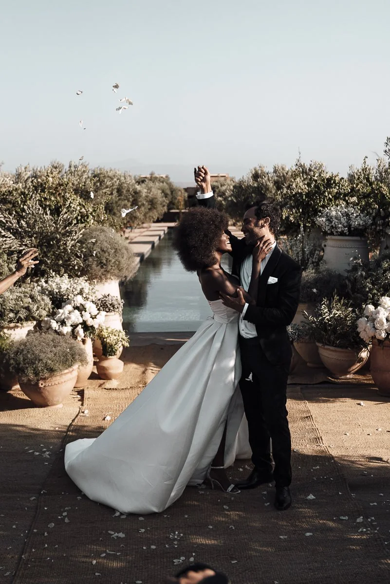 A bride and groom sharing a dance at their wedding, with the bride wearing a white gown and the groom in a black tuxedo, outdoors with trees and potted flowers.