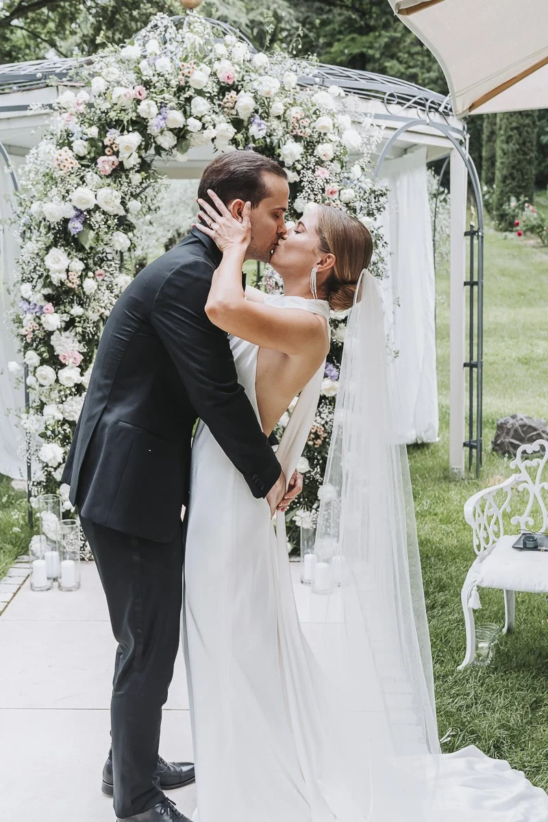 A newlywed couple kissing at their outdoor wedding ceremony with a floral arch and white furniture.