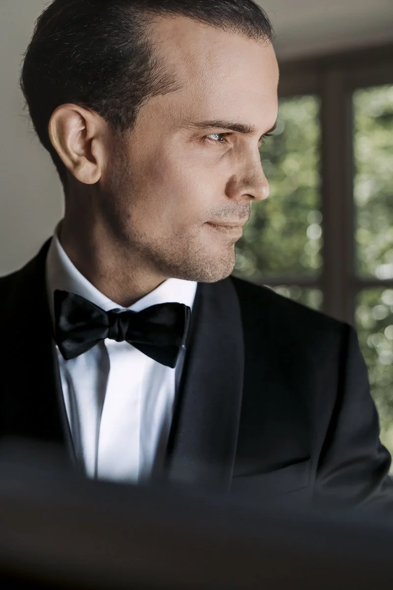A man dressed in a black tuxedo with a bowtie, looking to the right with a focused expression, standing indoors near a window with green foliage outside.