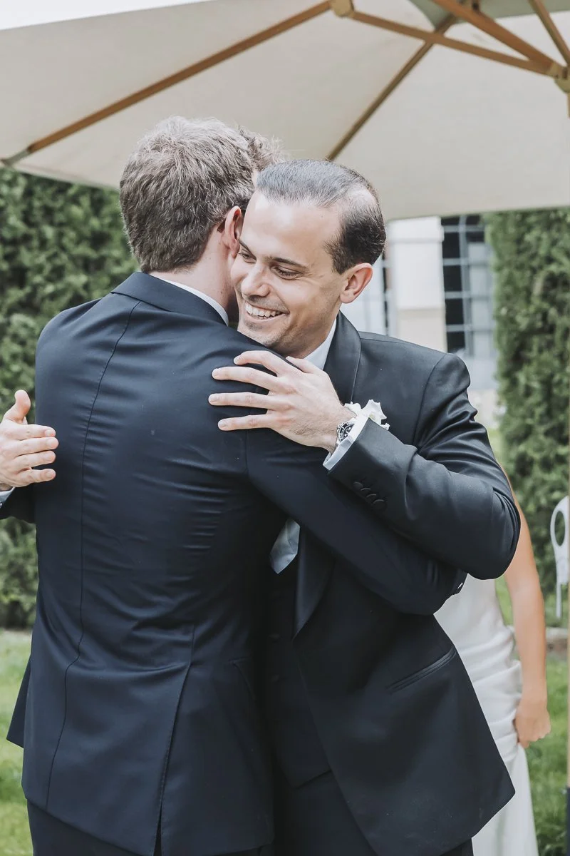 Two men in suits hugging and smiling at each other outdoors under a patio umbrella.