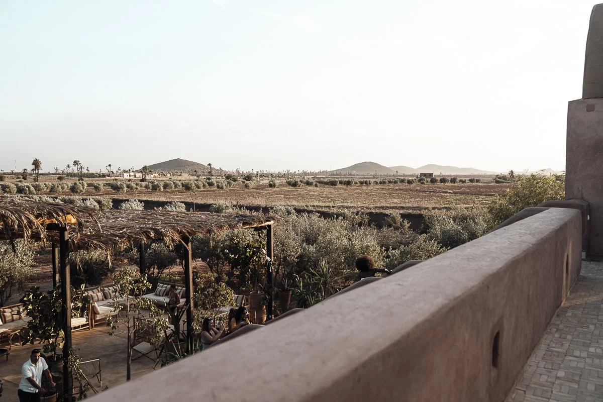 View from a balcony overlooking a desert landscape with sparse vegetation, including small bushes and palm trees. There is an outdoor seating area with tables and chairs, shaded by thatched roofs, and a few people are sitting and standing nearby.