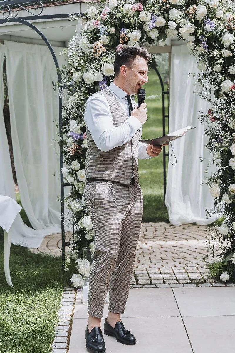 A man in a vintage-style suit giving a speech at a wedding ceremony outdoors, standing under a floral arch with white and pastel flowers, holding a book and a microphone.