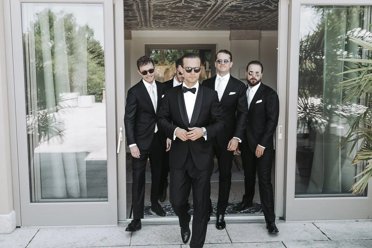 A group of five men dressed in black tuxedos, walking through a sliding glass door. Seven people are visible, with the man at the front wearing sunglasses, a bow tie, and a watch, leading the group.