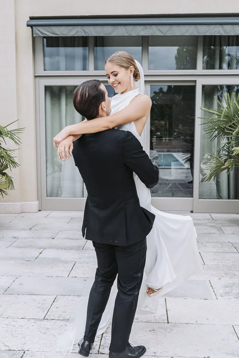 A bride and groom dancing outdoors on a patio, with the groom lifting the bride in his arms, smiling warmly at each other.