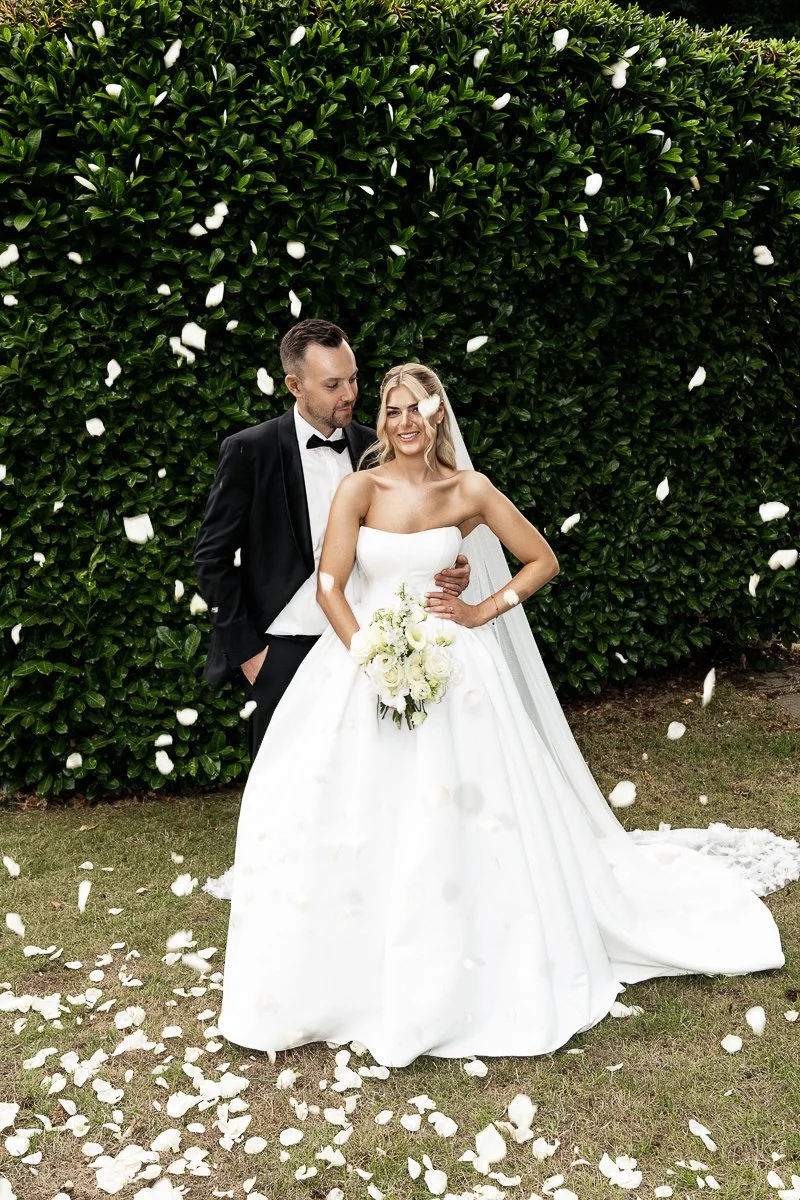 Brautpaar Laura und Lukas im Regen aus Rosenblättern vor grüner Hecke
