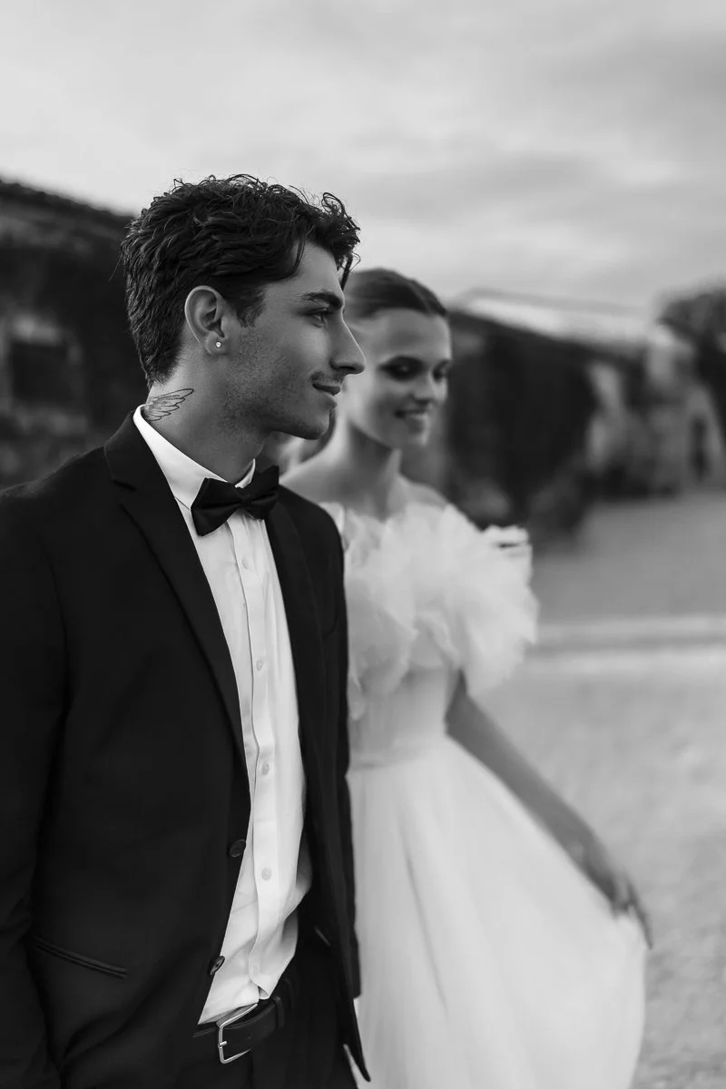 A black and white photo of a groom and bride on a beach, the groom wearing a tuxedo with a bow tie and the bride in a wedding dress. They are smiling and looking into the distance.