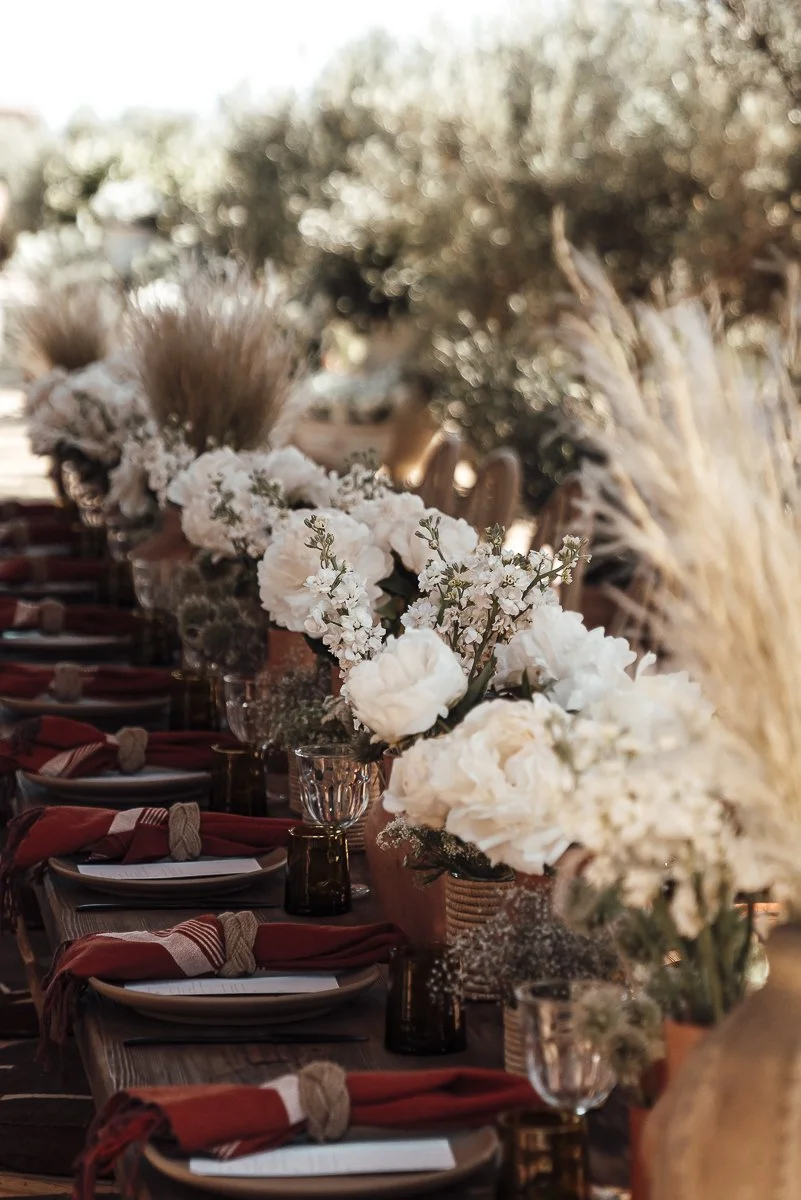 A beautifully set outdoor dining table decorated with white floral arrangements and brown glassware, with rust-colored napkins rolled around utensils.