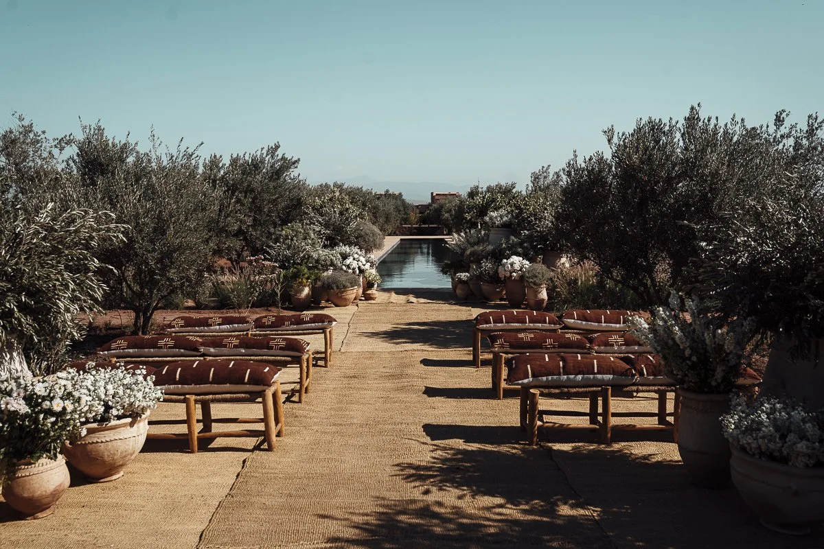 Outdoor patio with wooden benches, potted plants, and a swimming pool in the distance under a clear blue sky.