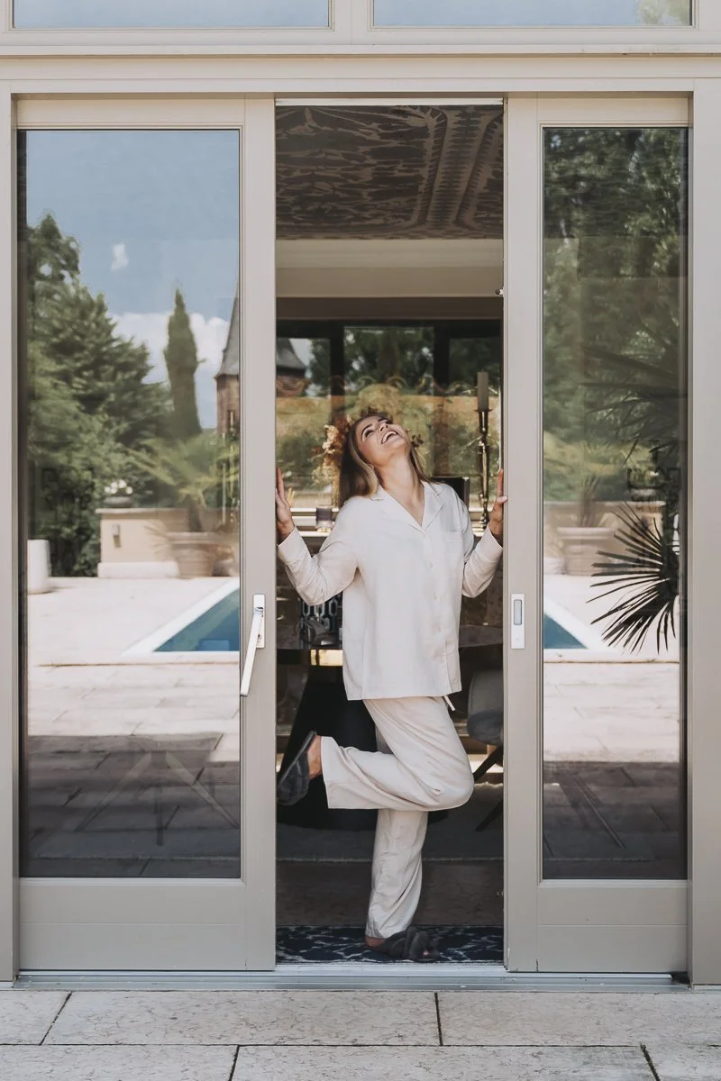 Woman in white pajamas smiling and posing playfully at sliding glass door, with outdoor pool and trees in background.