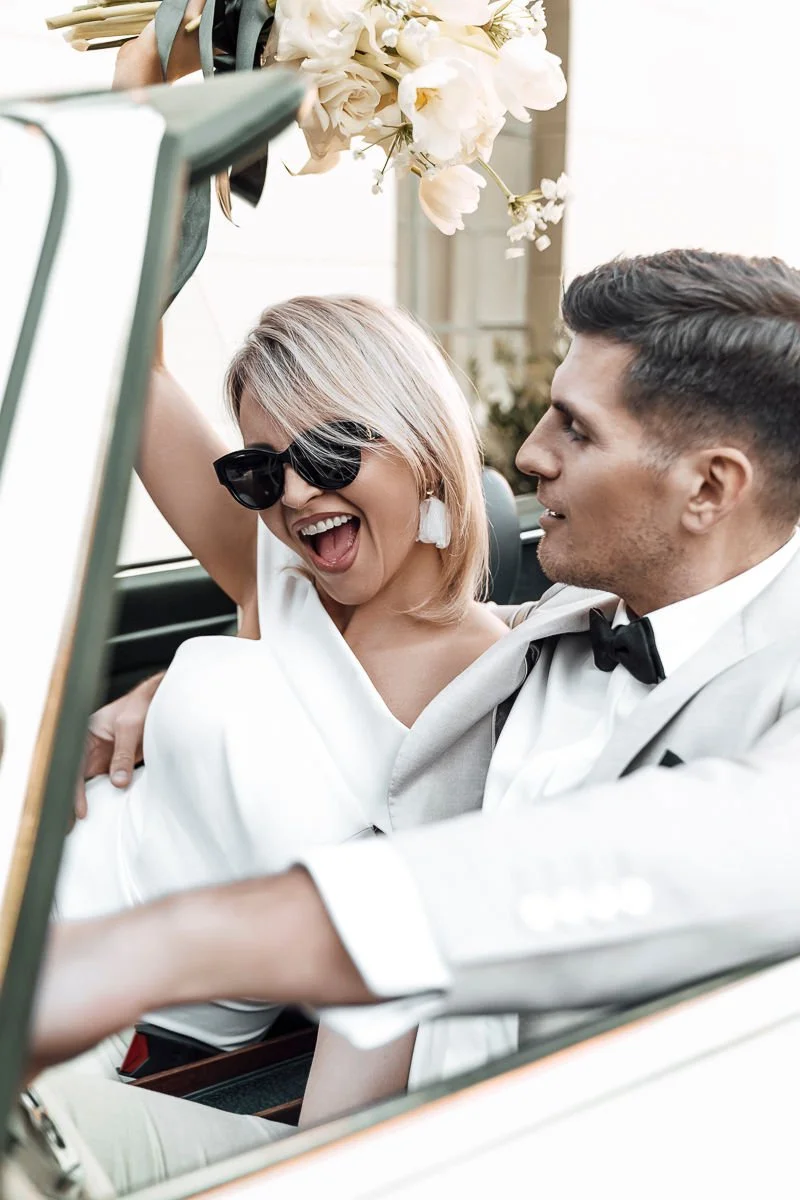 Lachende Braut mit Sonnenbrille und Bräutigam im Cabrio