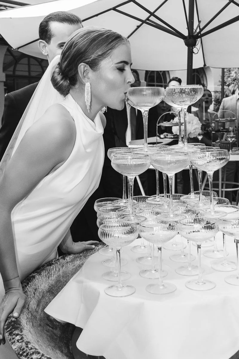 A woman in a white dress and wedding veil is leaning over a table, drinking from a champagne coupe glass placed on a pyramid of similar glasses, with a man in a black suit and other people in the background at a wedding reception under a large umbrel