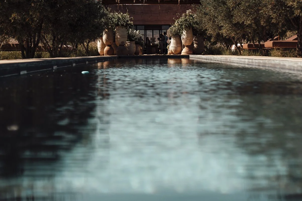A close-up, low angle view of a swimming pool reflecting the sky and trees, with a building and people in the background.