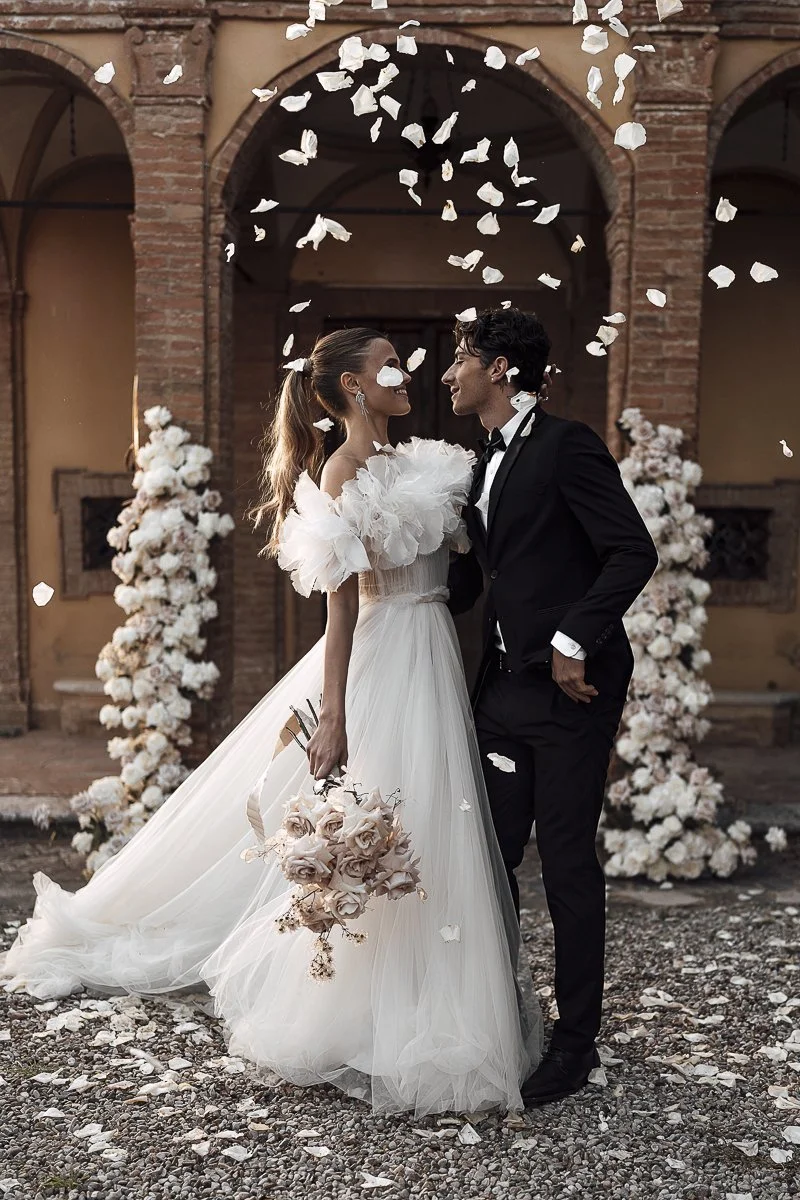 A bride and groom in wedding attire standing close with their faces almost touching, smiling at each other surrounded by falling flower petals, outside in front of an arched brick structure decorated with large white floral arrangements.