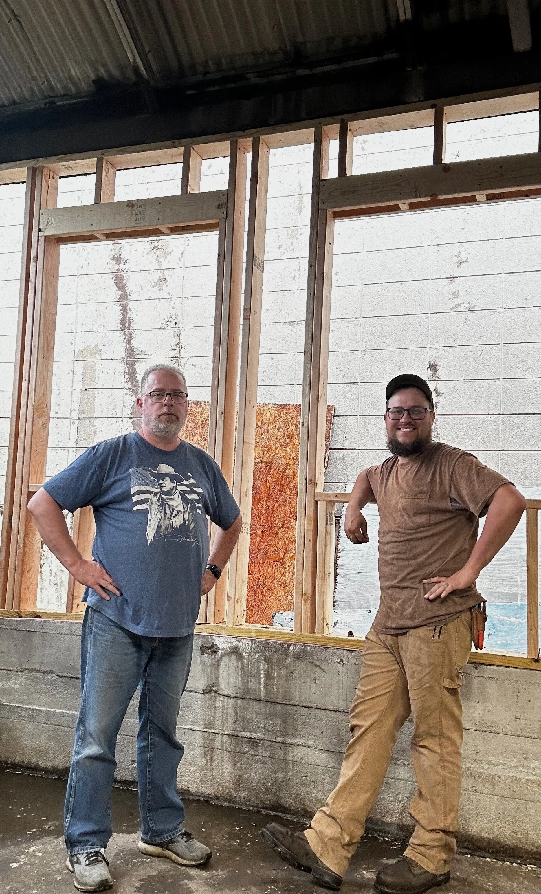 Two men standing inside a building under construction, with exposed wooden framing and concrete foundation.
