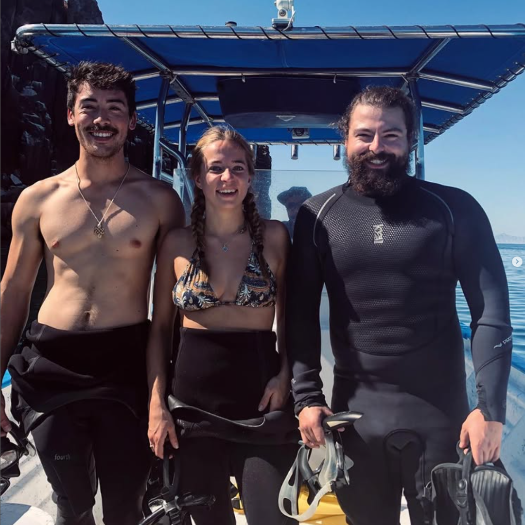 Three people smiling on a boat, dressed in wetsuits and swimwear, with the ocean and blue sky in the background.