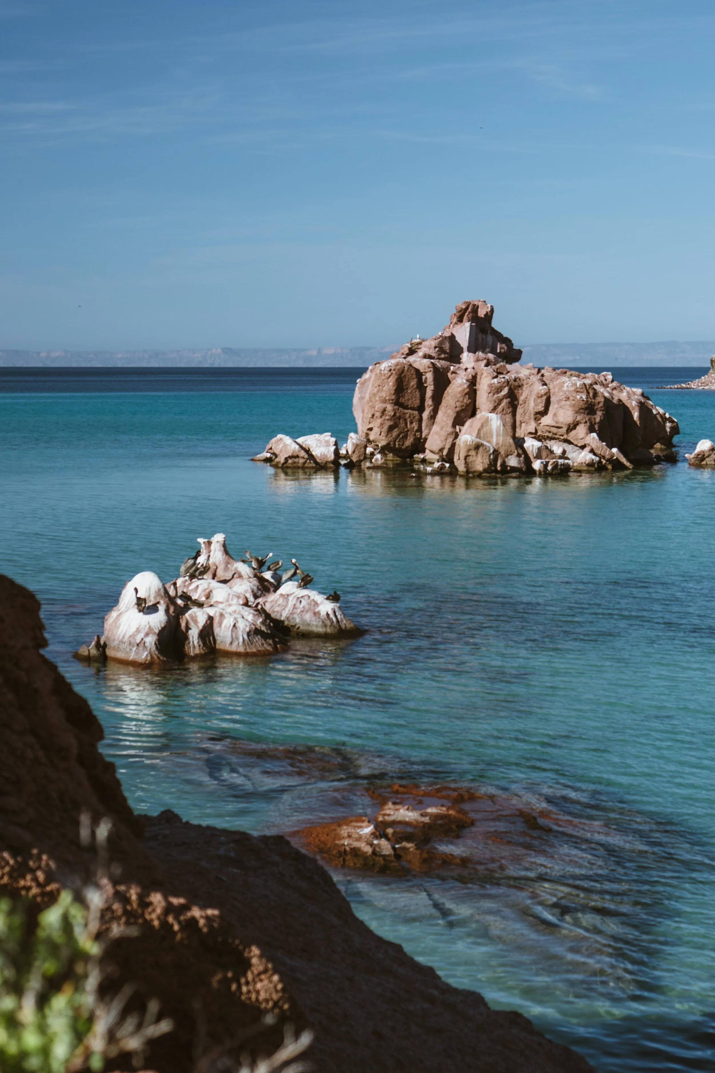 A coastal scene featuring large rocks and boulders in clear turquoise water, with smaller rocks and seabirds perched on them. In the background, a larger rocky formation rises from the water, under a blue sky with light clouds.