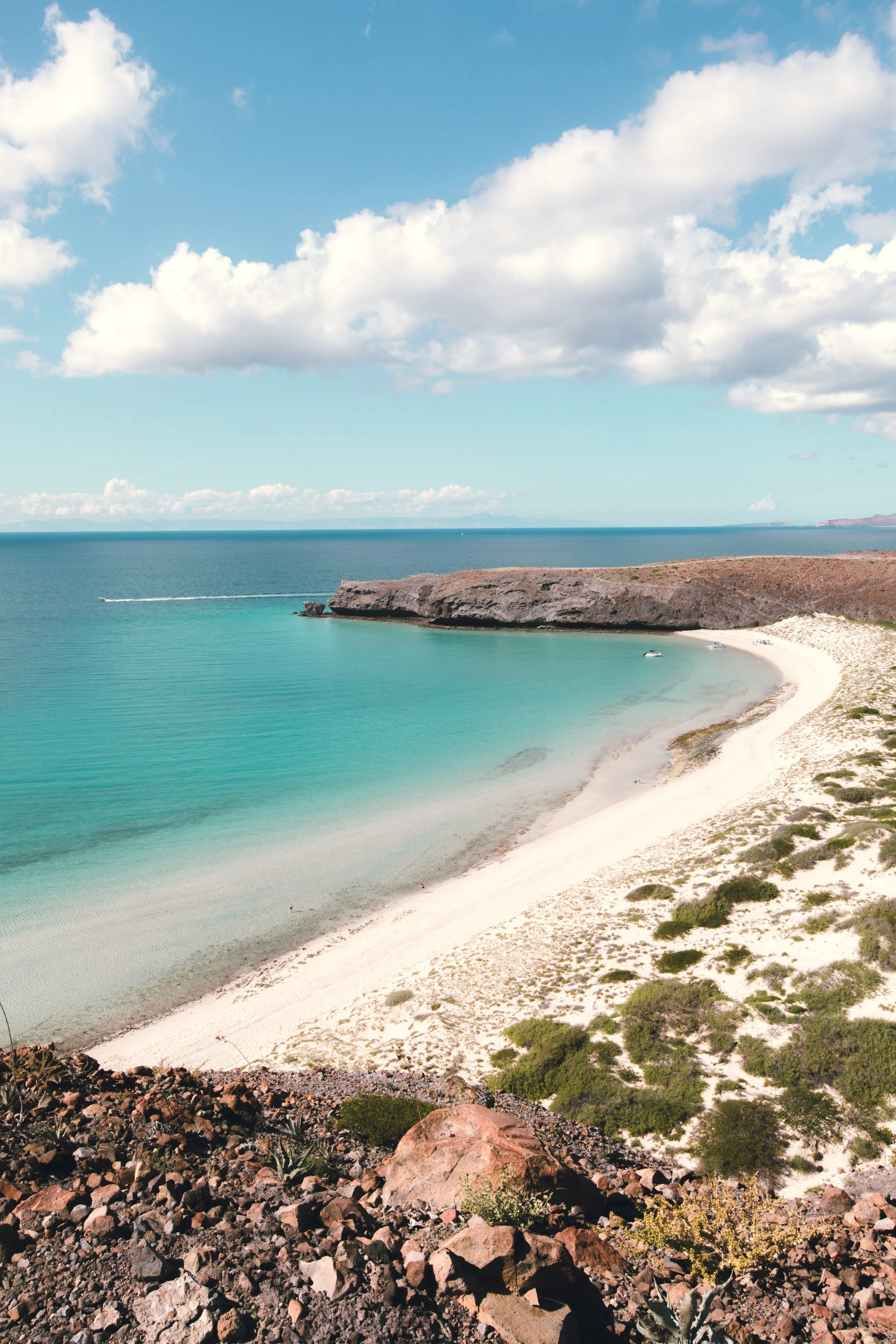 A beach with white sand, turquoise water, and a rocky cliff in the distance under a partly cloudy sky.