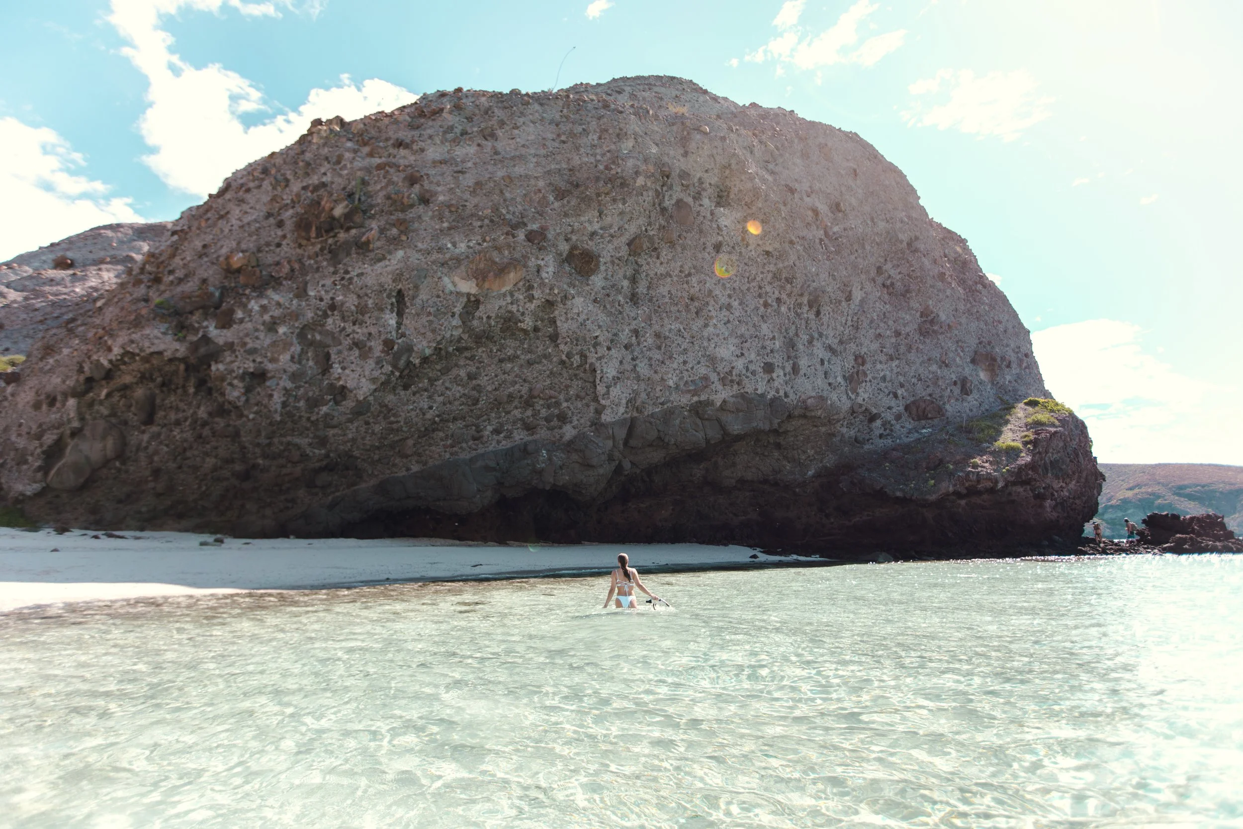 A woman in a white swimsuit standing in shallow clear water near a sandy beach, facing a massive rocky cliff under a partly cloudy sky.