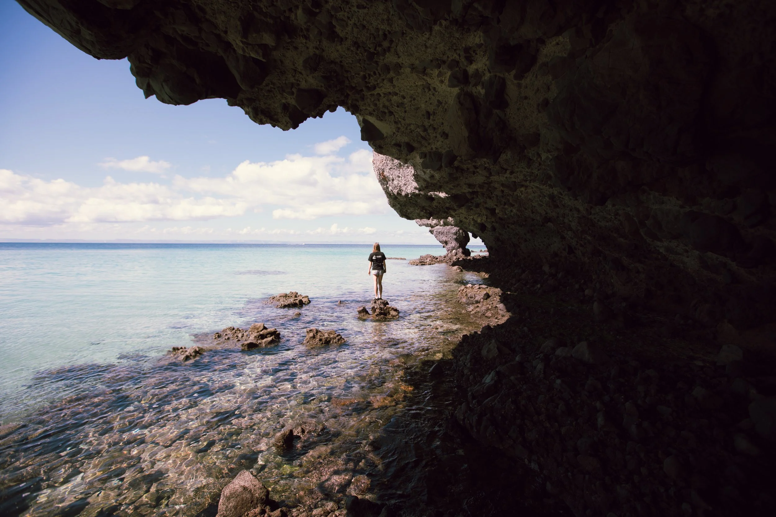 A person hiking along the shoreline of a beach, with rocky cliffs forming a cave-like structure overhead, facing the ocean on a partly cloudy day.