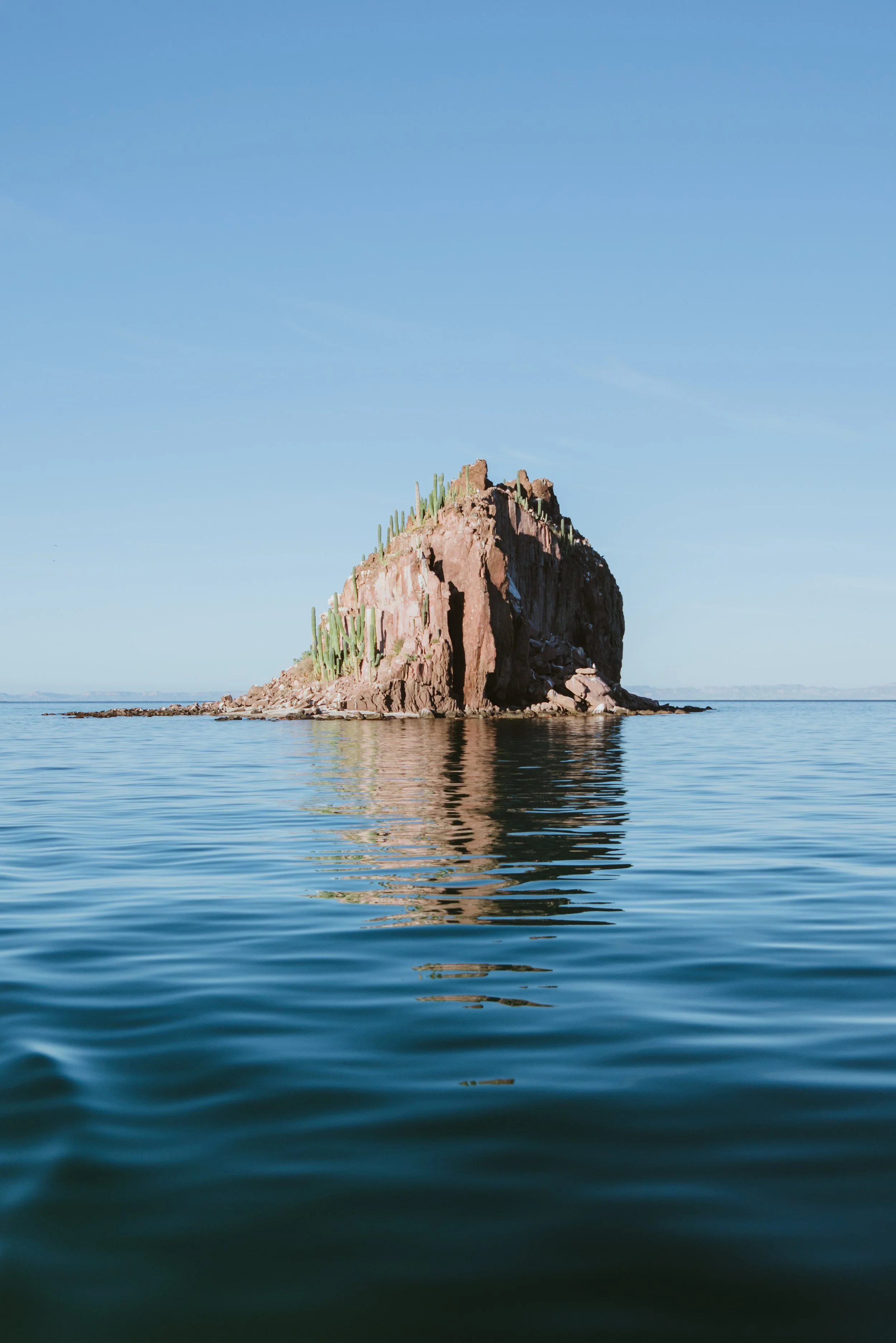 A large rocky island with cactus plants on top, reflected in calm water under a clear blue sky.