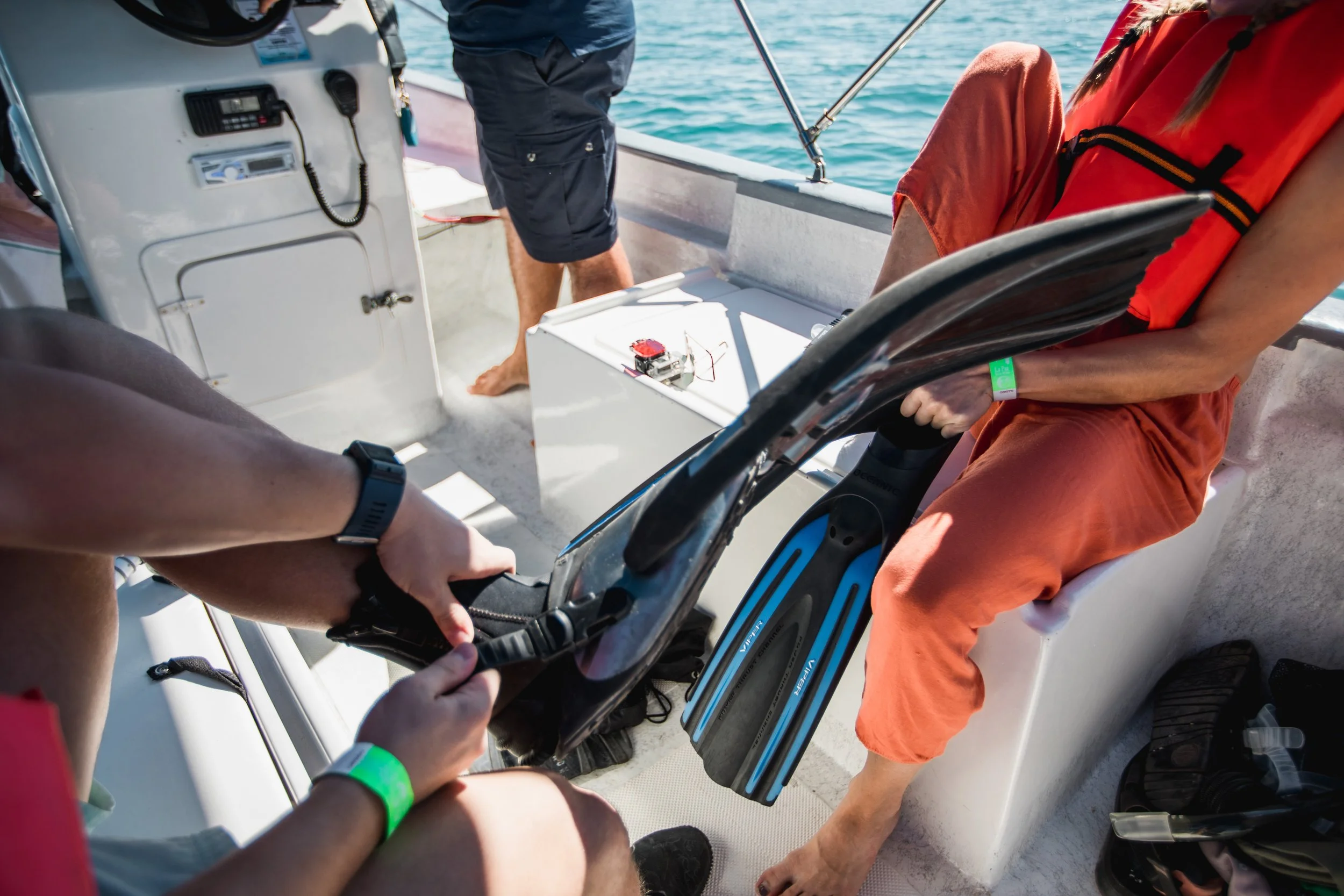 People on a boat preparing for a dive, with one person sitting and another holding a scuba diving regulator, and a third person standing.”}