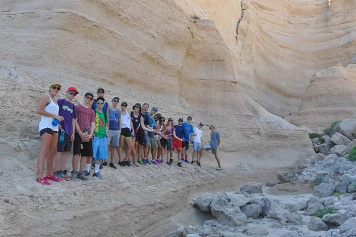 A group of people standing on a narrow ledge beside a large beige rock formation with a riverbed below.