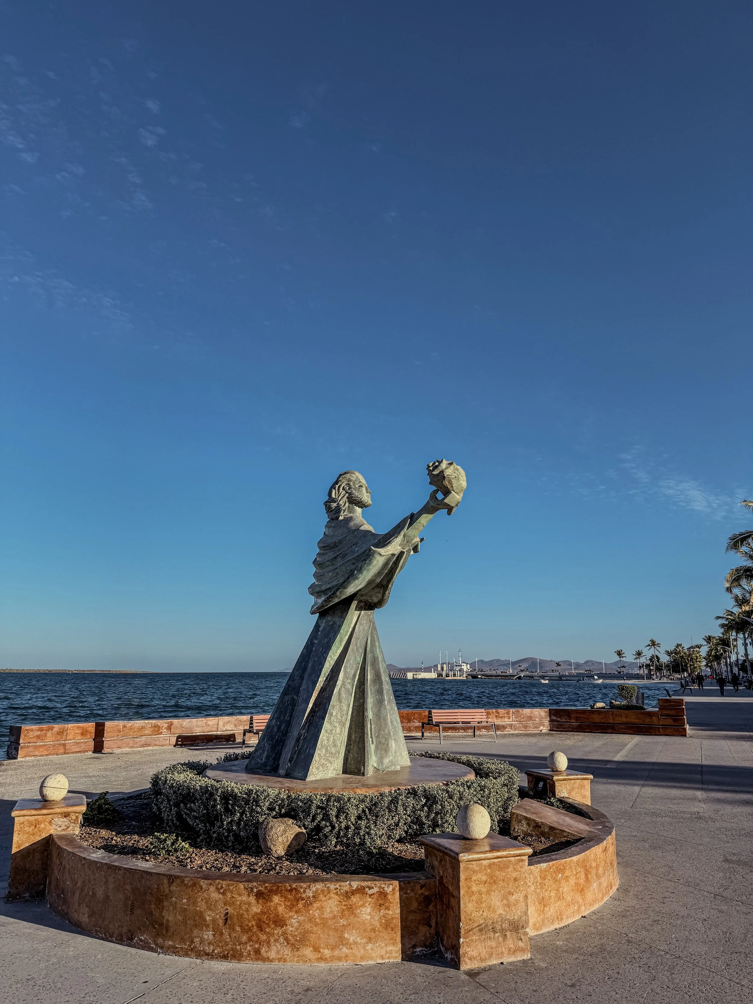 Statue of a man holding a globe in a promenade near the water and palm trees under a blue sky.