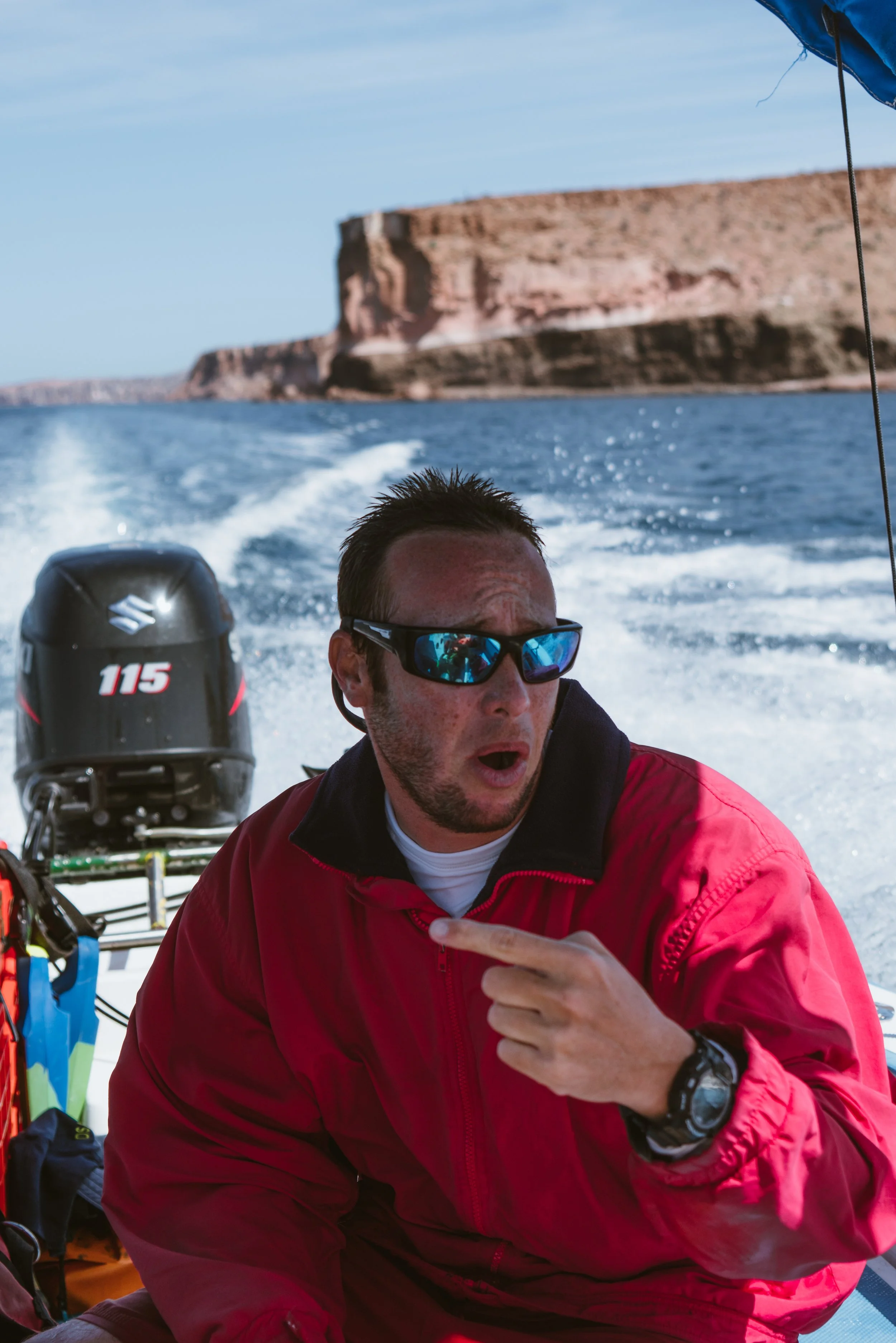 Man wearing sunglasses in a red jacket pointing to his chest on a boat, with water and distant cliffs in the background.